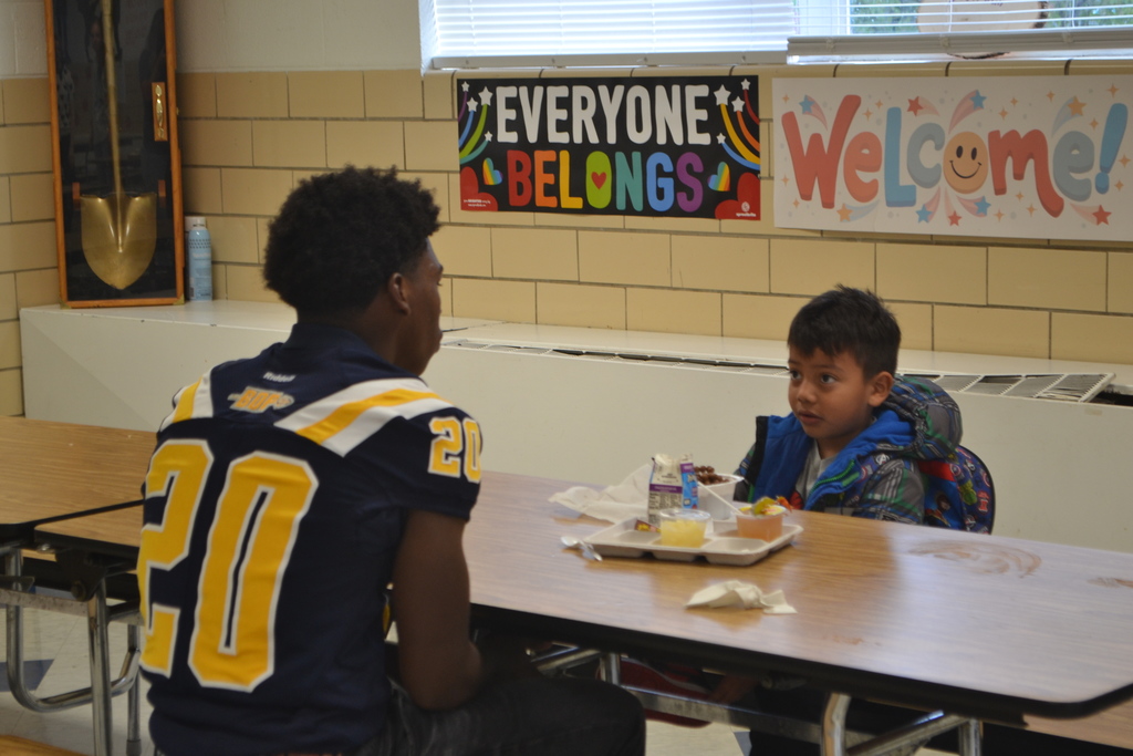 A football player (20) is sitting in the school cafeteria talking to a younger student while he eats breakfast.  EVERYONE BELONGS  WELCOME is on the sign in the background.