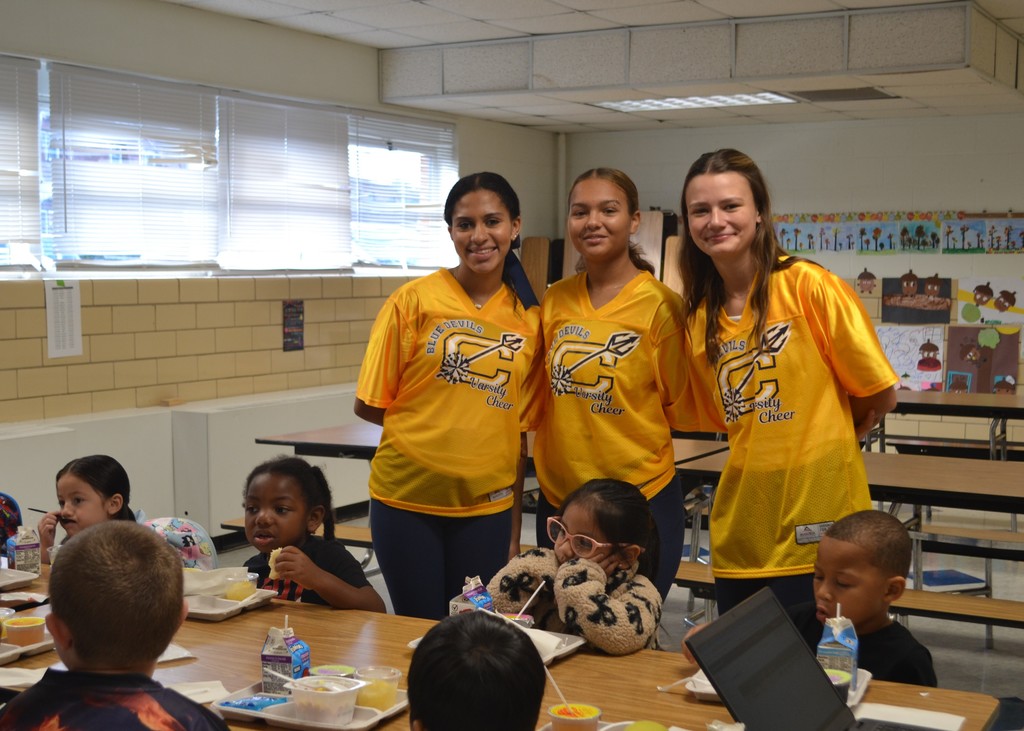 Three cheerleaders in yellow shirts smile behind a table of kids eating breakfast.