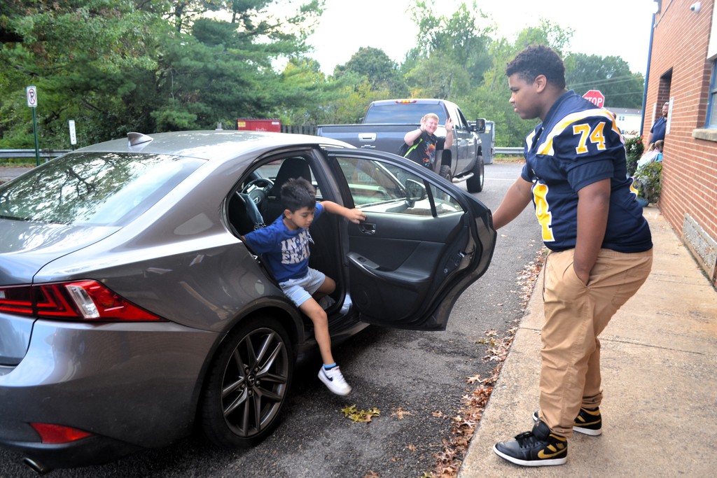 A football player (74) opens the car door to a student getting out.  Another student waves in the background.  (School drop off line)