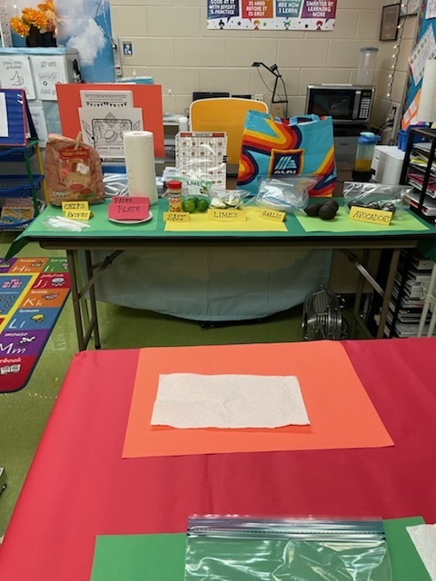 Classroom table setup with labeled supplies for making guacamole, including avocados, limes, garlic, salt, chips, plates, and utensils, neatly organized with colorful signs.
