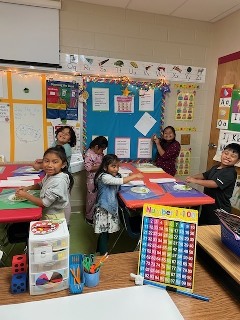 First graders around a classroom table actively preparing guacamole. The table is covered in colorful mats and the students are smiling as they work together.