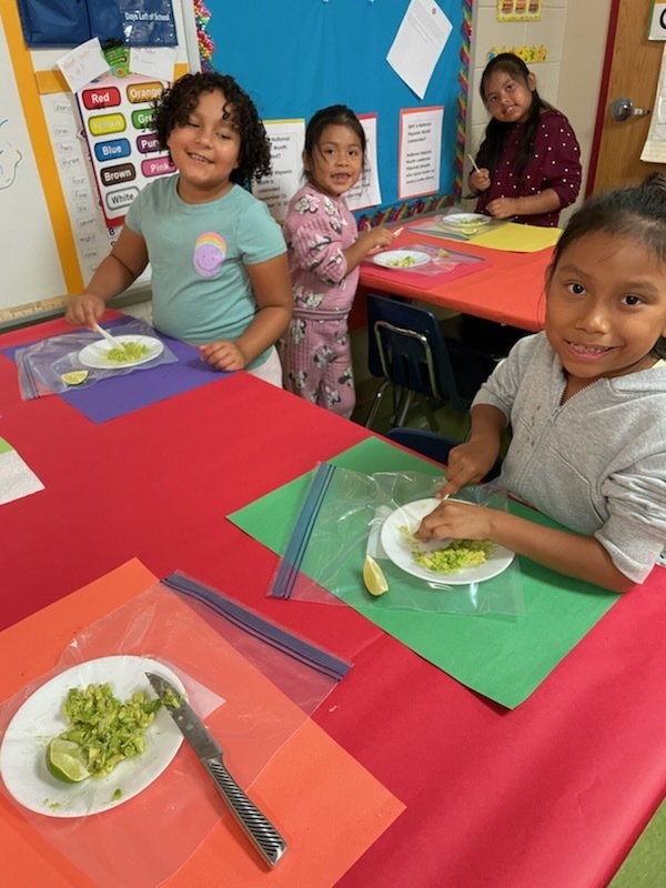 Small group of first graders smiling at a table while mashing avocados on plates. They are using plastic bags and utensils as part of a guacamole-making activity.