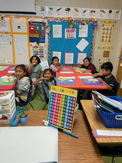 Group of first graders seated at a table enjoying plates of guacamole with lime wedges and tortilla chips. The students are smiling as they taste their finished recipe.