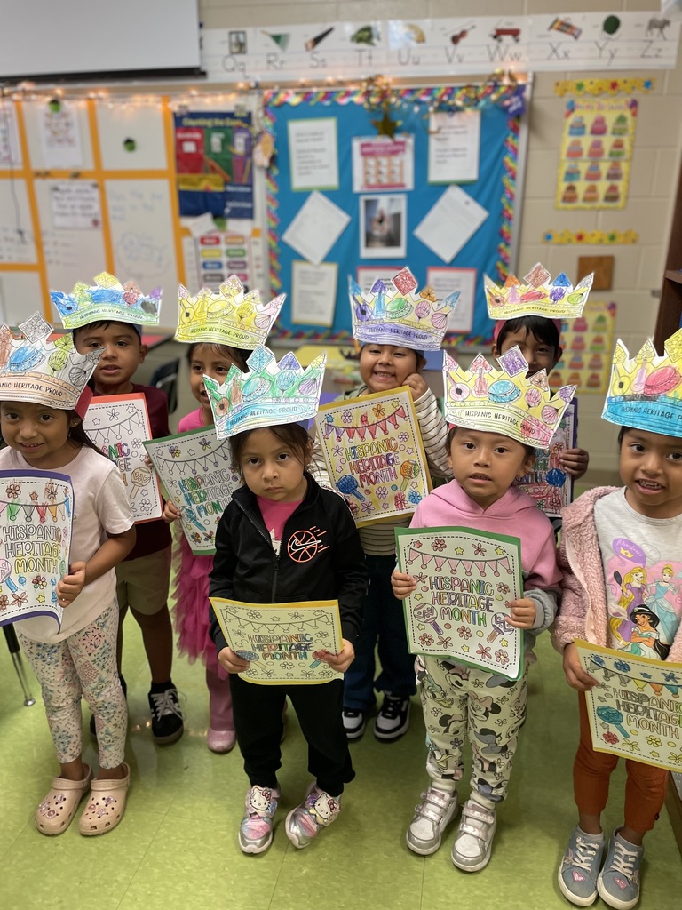 Group of kindergarten students wearing brightly colored paper crowns that read “Hispanic Heritage Proud.” They hold up their decorated papers for Hispanic Heritage Month while smiling in the classroom.