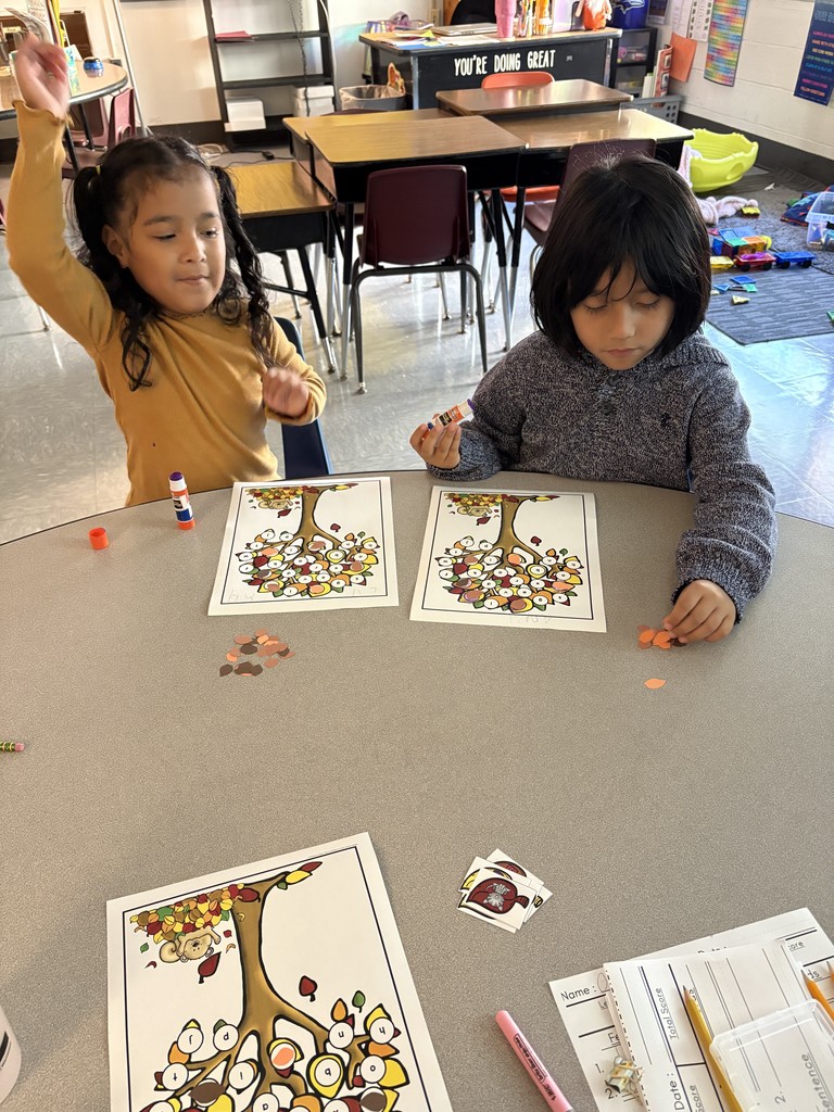 Two young children sit at a round classroom table working on a fall-themed craft. They each have a paper tree in front of them with leaves labeled with letters, and they are gluing small paper leaves onto the branches. One child is holding up their hand excitedly, while the other is focused on gluing.