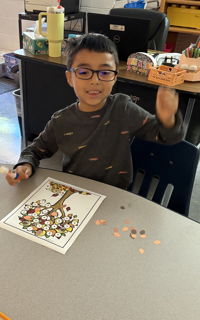A young child wearing glasses sits at a table in a classroom, working on the same fall tree activity. They are holding a glue stick and smiling, with small cut-out leaves scattered on the table.