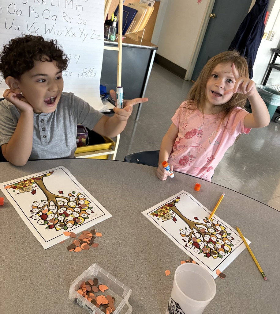 Two young children sit together at a table with their fall tree activity sheets. Both are smiling and gesturing playfully toward the camera. The table has glue sticks, pencils, and paper leaves spread out for the project.