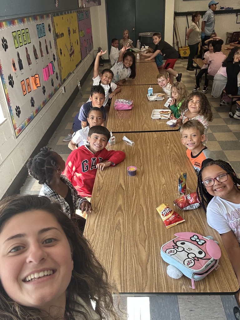 Ms. Powers poses with a long line of students at a cafeteria table. The kids beam at the camera, holding lunches and snacks, with colorful wall decorations in the background.