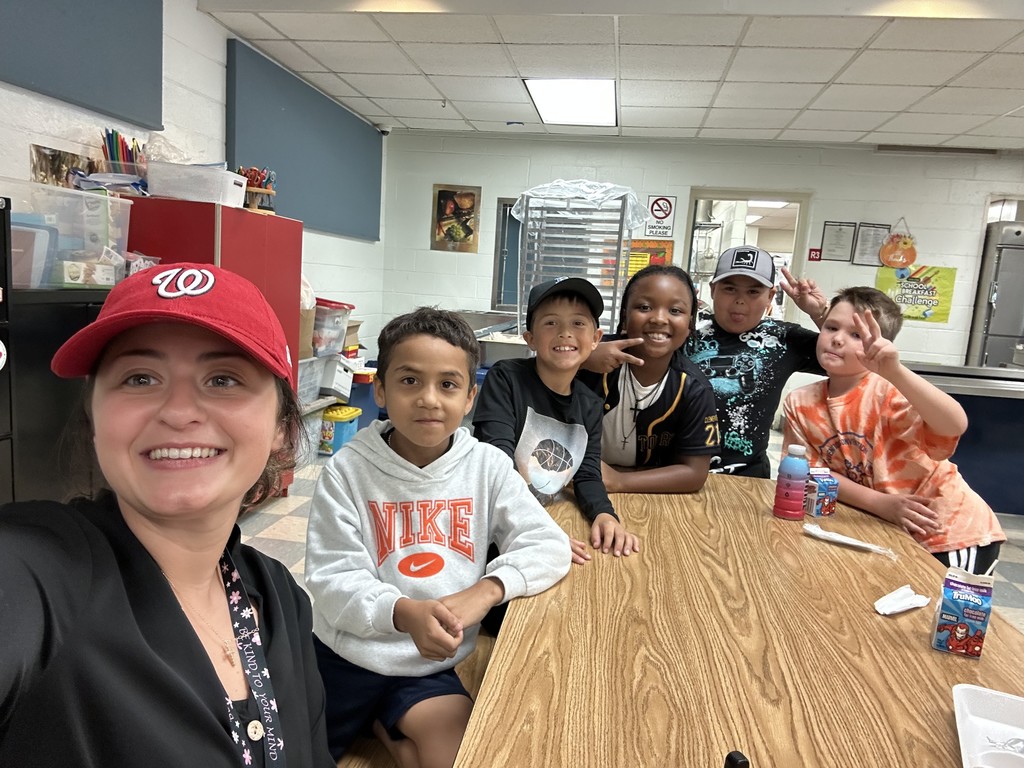 Ms. Powers, wearing a red hat, smiles with a small group of students at a cafeteria table. The students grin and make peace signs, with their milk cartons and snacks on the table.