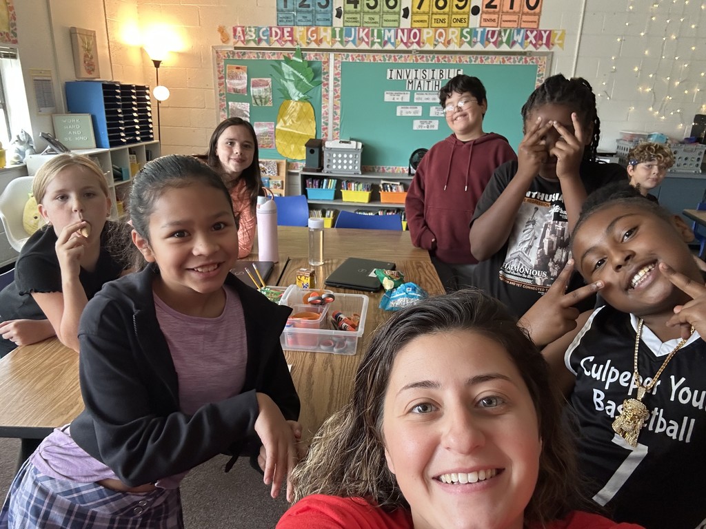 Ms. Powers takes a selfie with a group of older elementary students in a classroom. The kids smile, laugh, and make playful hand signs while snacks and school supplies are on the table behind them.