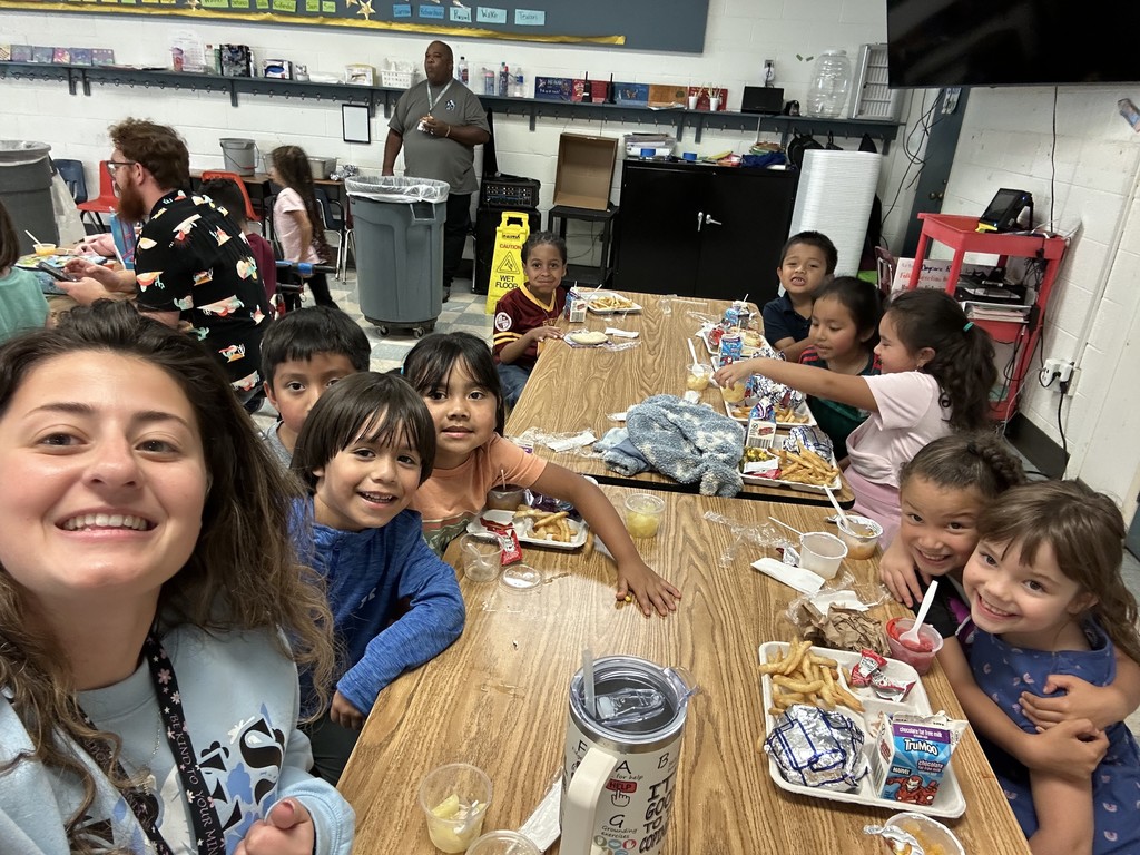 Ms. Powers smiles at a cafeteria table with a group of young elementary students. The children are eating lunch trays filled with fries, fruit cups, and milk, while smiling and leaning in for the photo.