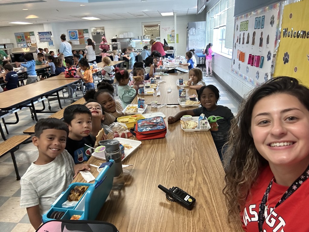 Ms. Powers sits at a long cafeteria table with young students enjoying lunch. The kids smile brightly at the camera, with trays of school lunch and milk cartons in front of them. The cafeteria is filled with students in the background.