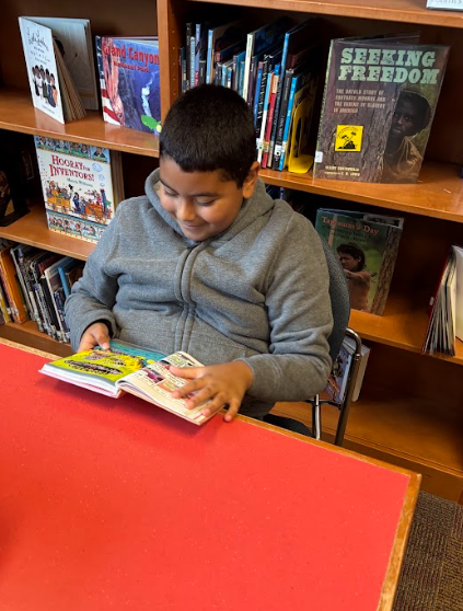 A student wearing a gray hoodie is smiling while reading a colorful graphic novel at a red library table.