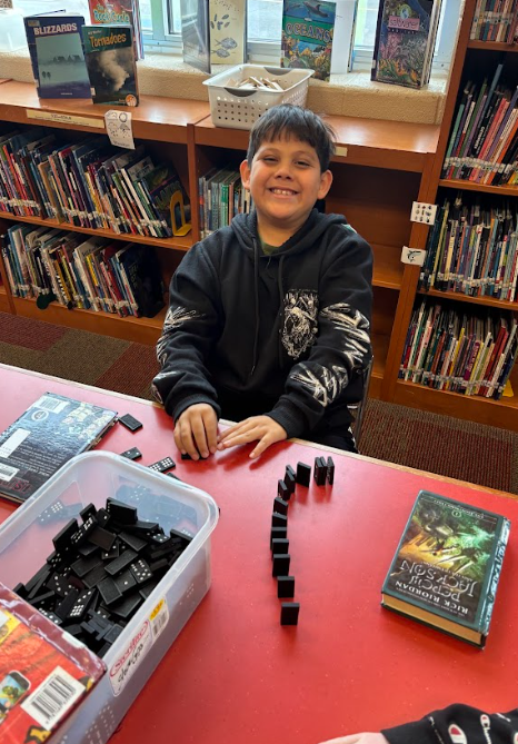 A student in a black hoodie is smiling while sitting at a red library table, arranging dominoes into a curved line with books visible behind him.