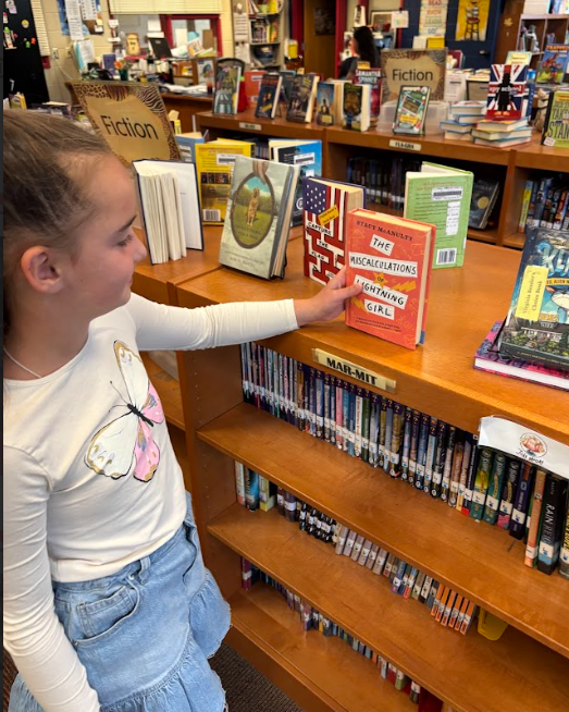 A student wearing a white shirt with a butterfly design is pointing to a book titled The Miscalculations of Lightning Girl on display in the library.