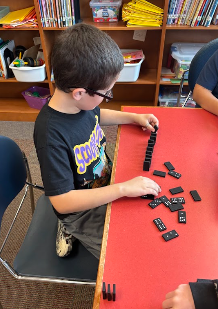 A student in a black Scooby-Doo shirt is arranging black dominoes into a line on a red library table.