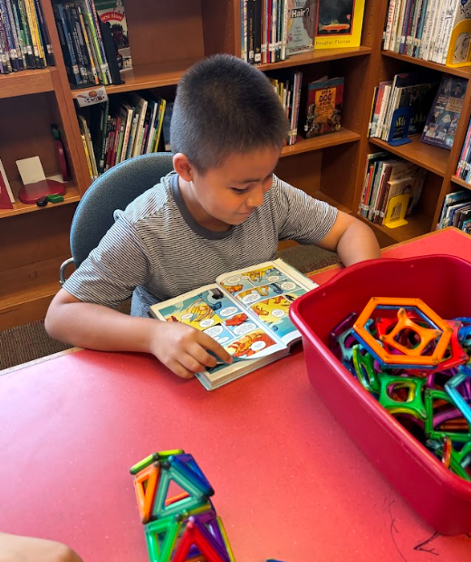 A student in a striped shirt is focused on reading a comic book at a red library table with colorful magnetic building tiles beside him.