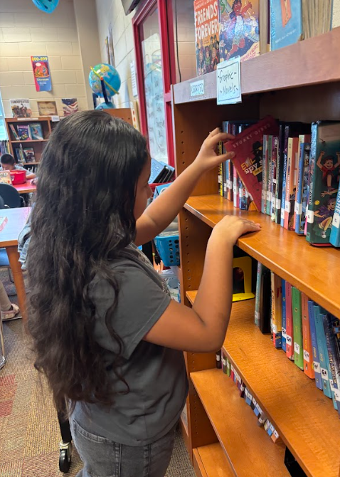 A student with long dark hair is placing a book on a wooden library shelf labeled “Graphic Novels.”