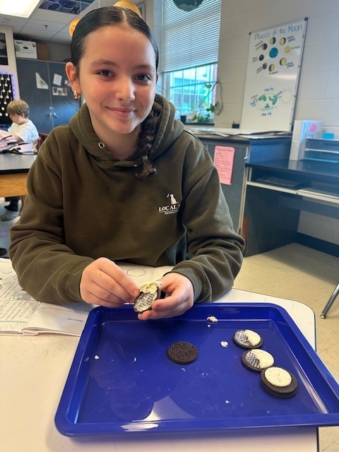 A student in a classroom smiles while separating the cream from sandwich cookies on a blue tray. A moon phases poster is visible behind them.