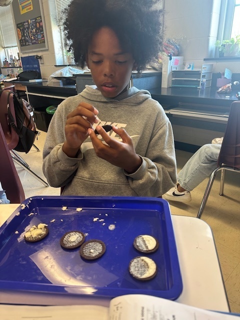 A young person in a classroom decorates cookies with icing, seated at a table with a blue tray of cookies. They appear focused and engaged.