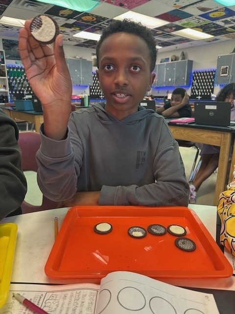 A student in a classroom holds up a cookie with its cream side facing forward. Several cookies lie on an orange tray beneath him. The classroom has a warm, educational atmosphere.