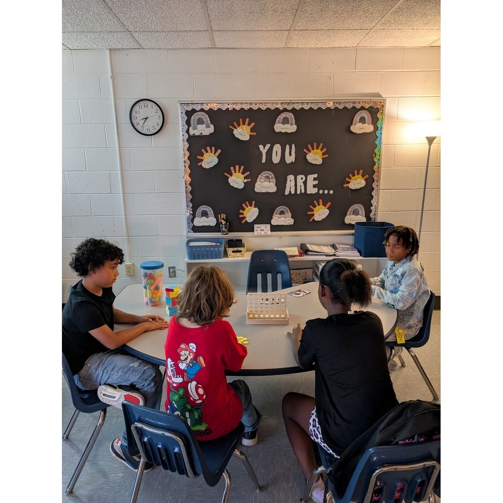 Four children sit around a classroom table playing educational games, with a bulletin board behind them reading “YOU ARE…” decorated with rainbows and suns.