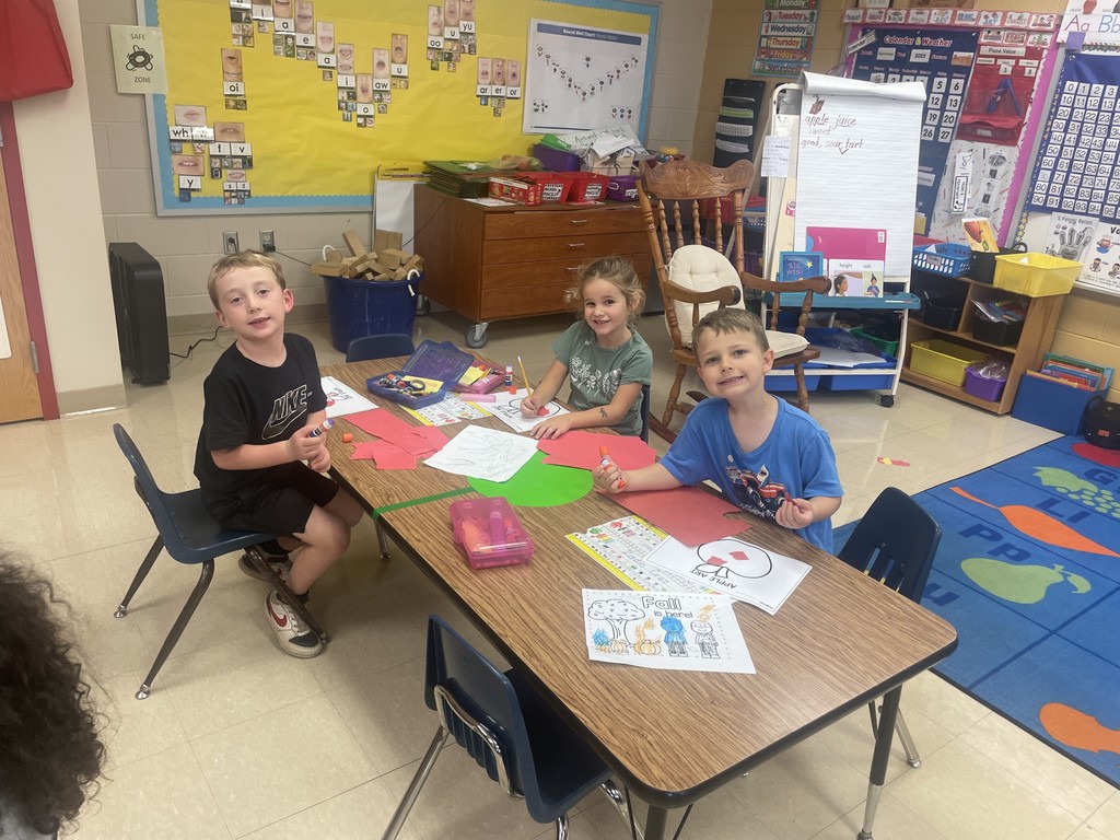 Three young students sit at a classroom table smiling as they work on apple-themed crafts. The children are using glue sticks, crayons, and colored paper shaped like apples. A bright bulletin board with phonics cards and a “Safe Zone” sign is visible behind them.