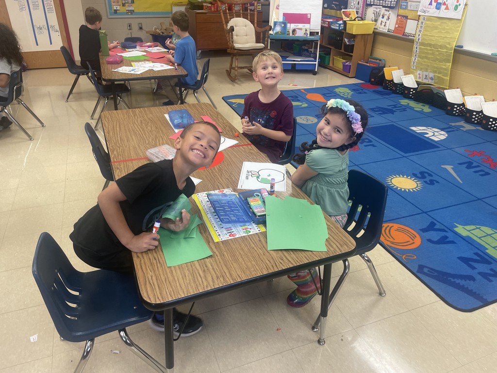 Three smiling students sit together at a table working on apple art projects. The children use green and red paper and glue sticks. The classroom carpet features colorful apples and letters, and other classmates can be seen working in the background.