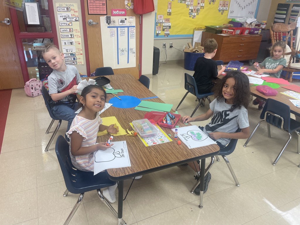 Four students sit around a table creating apple crafts with crayons and glue. Two children in the front smile at the camera, showing their work with apple outlines. The classroom background includes a yellow word-wall bulletin board and a small rocking chair near the corner.