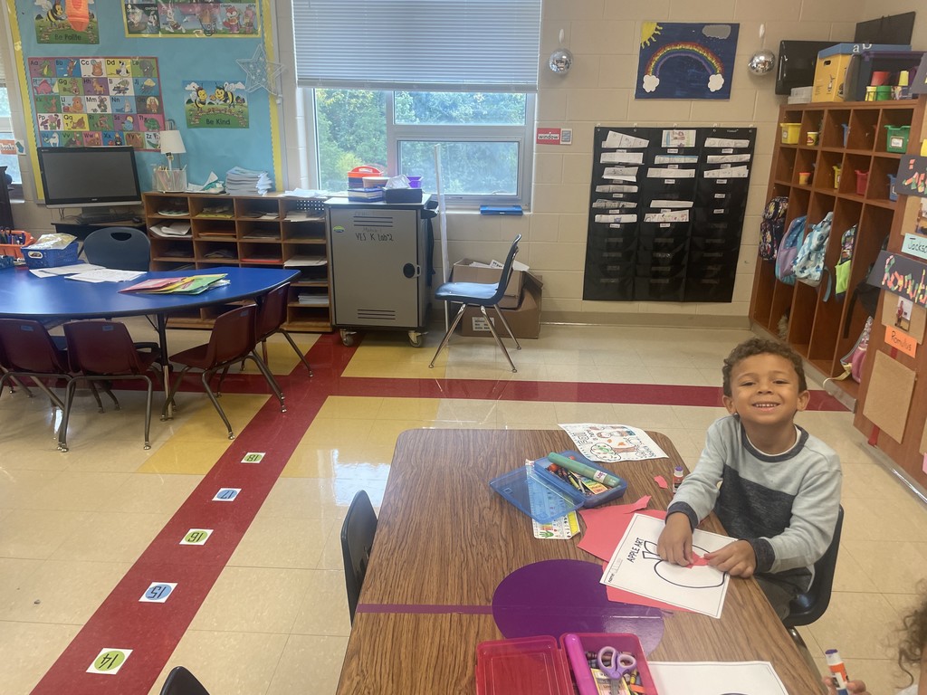 A smiling student sits at a classroom table holding a glue stick and working on an apple craft. The student’s table is covered with crayons, scissors, and colored paper. Behind him are cubbies, backpacks, and a bright classroom wall decorated with a rainbow painting and bee-themed posters.