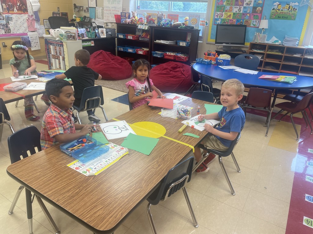 Four students sit at a table making apple crafts using paper, markers, and glue. The children are smiling and engaged in their work. Behind them, the classroom displays colorful posters, a “Be Kind” bulletin board, and beanbag chairs in a cozy reading corner.