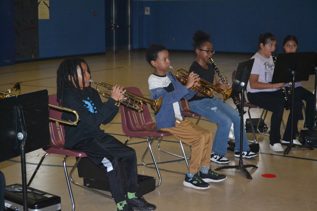 Five students in chairs with music stands playing their instruments at practice in the school gym.