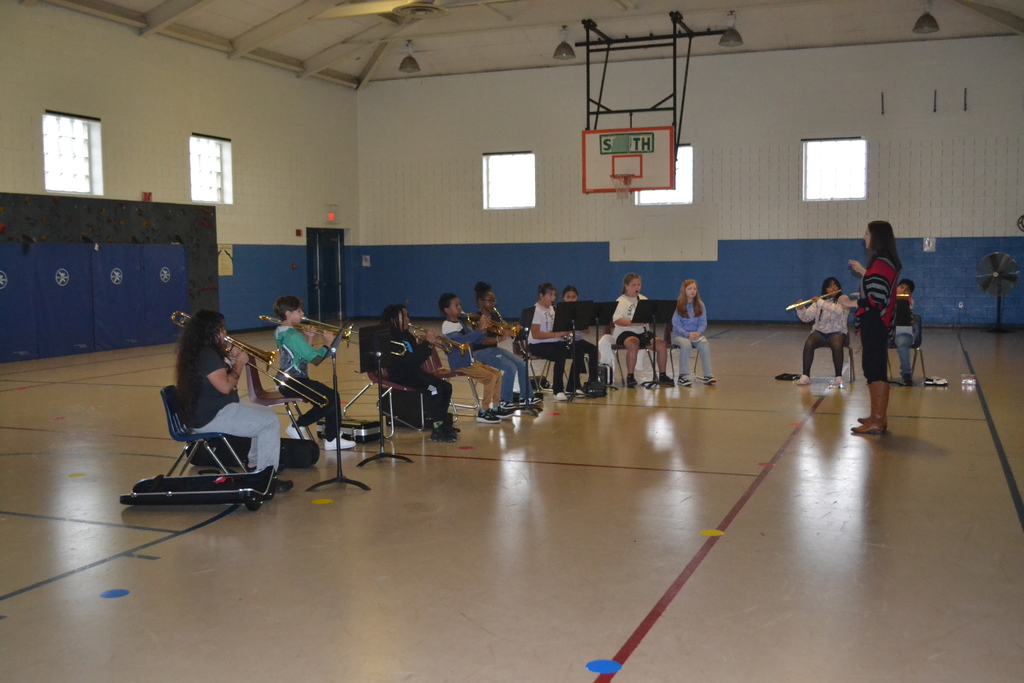 Director and students in chairs with music stands playing their instruments at practice in the school gym.