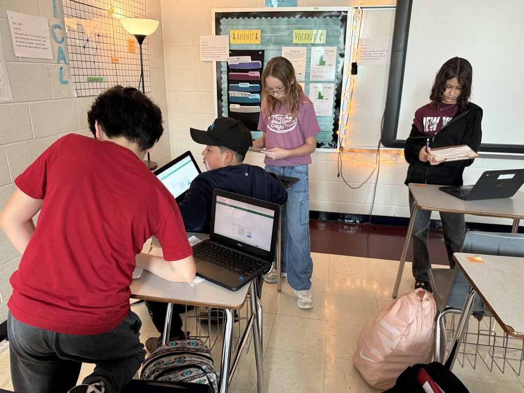Four students work collaboratively in a classroom, using laptops and notebooks. The room is decorated with learning charts, creating a focused atmosphere.