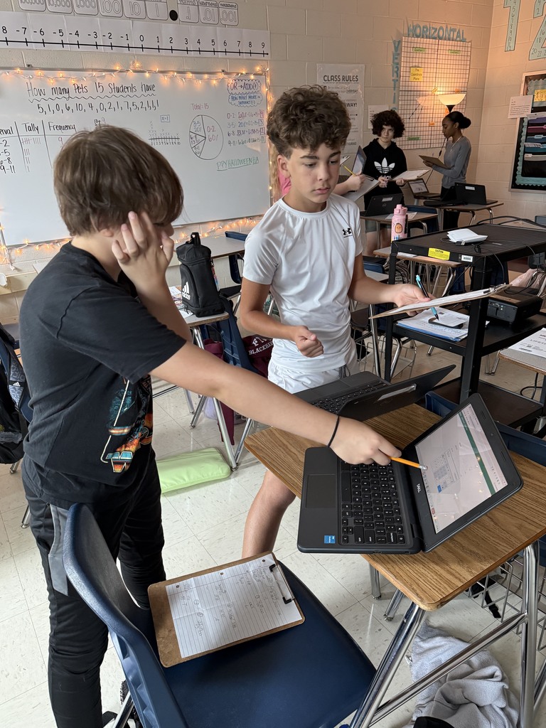 Two students collaborate in a classroom, one pointing at a laptop screen with a pencil, surrounded by desks and educational charts on the walls.