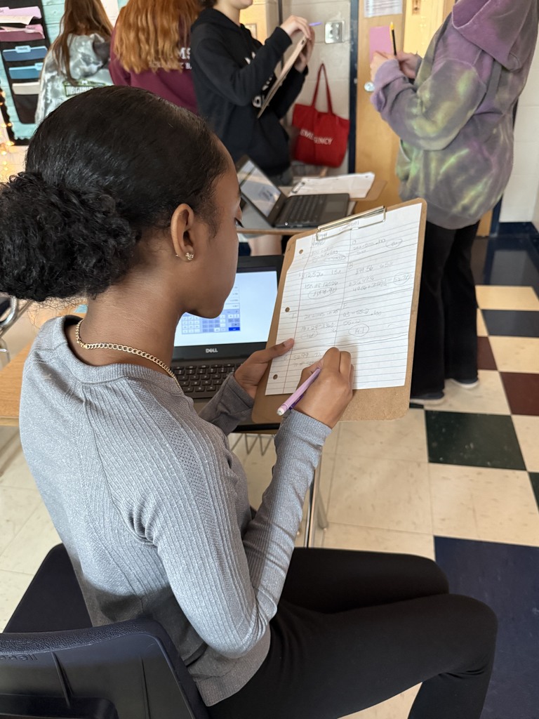 A student in a gray sweater sits in a classroom, holding a clipboard and writing notes. A laptop is open on the desk, and other students are in the background.