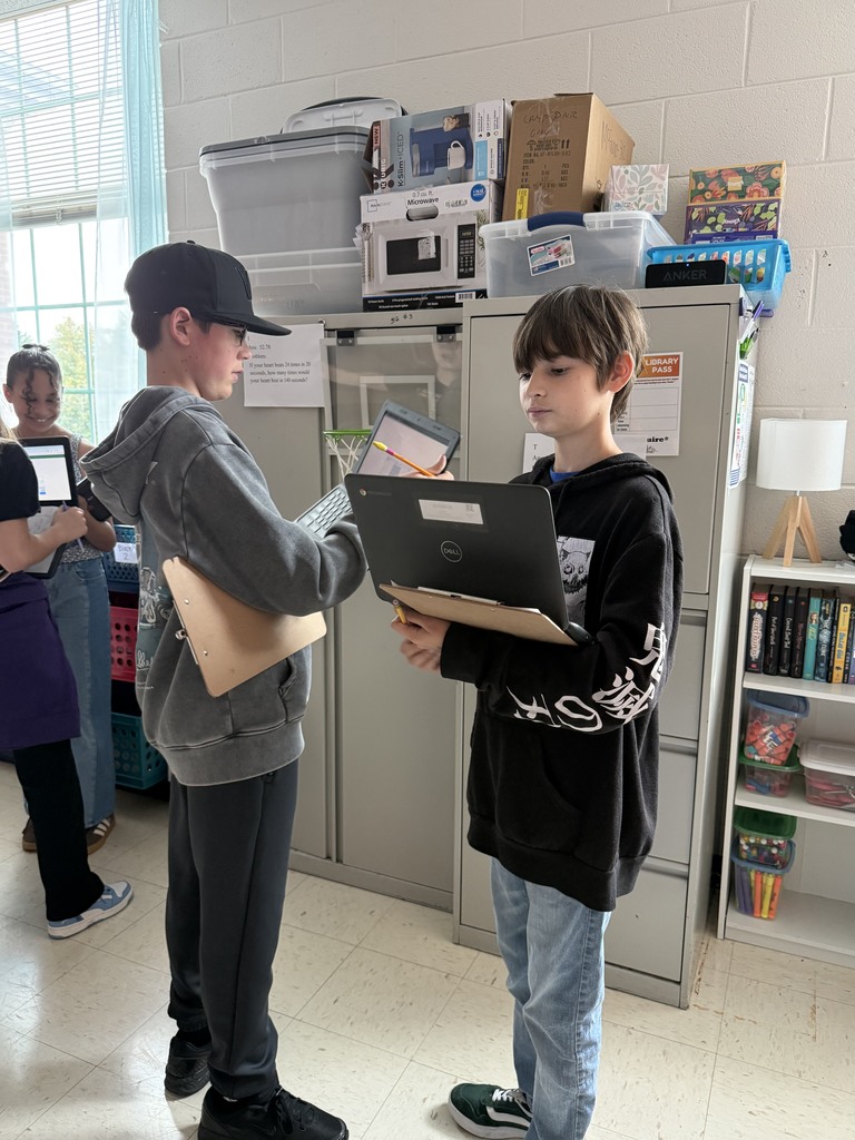 Two boys stand in a classroom with clipboards and laptops, engaged in discussion. A girl is in the background. Shelves and bins are visible.