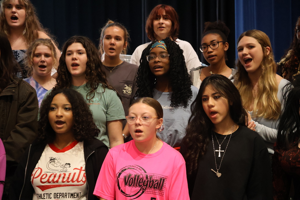 A group of high school students are singing together on stage. They are standing in rows, facing forward with their mouths open mid-song. The students wear a variety of casual clothing, including graphic t-shirts, sweatshirts, and glasses. The background is dark, drawing focus to the performers.