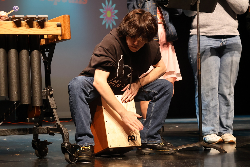 A student is performing on stage, sitting and playing a wooden cajón drum with one hand while leaning forward. They are wearing jeans, a black hoodie, and black shoes with yellow laces. Other musicians and instruments, including a marimba and music stands, are partially visible around them.