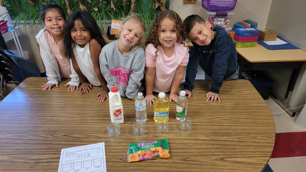 Five smiling young students stand behind a classroom table displaying materials for a science experiment. On the table are four jars, a bottle of milk, a bottle of water, vegetable oil, vinegar, and a bag of Brach’s pumpkin candies. A worksheet labeled “Dissolving Pumpkins Science Experiment” is visible in the foreground.