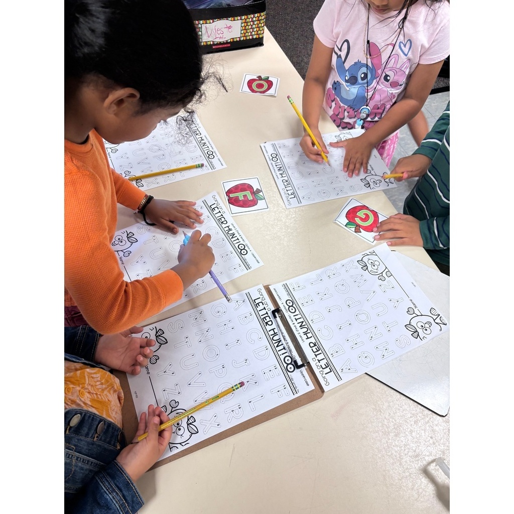 Four students at a table working on alphabet letter hunt sheets with pencils.