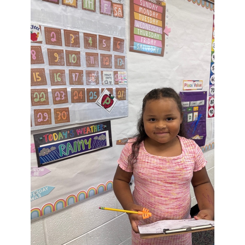 A child in a pink dress stands with a pencil and paper in front of a classroom calendar and weather chart.