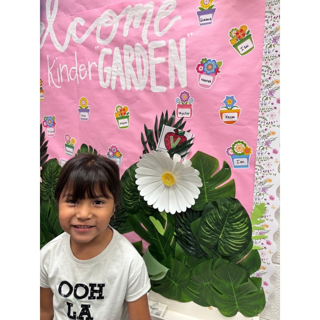 A child stands in front of a pink “Welcome to Kindergarten” display with flowers labeled with student names.