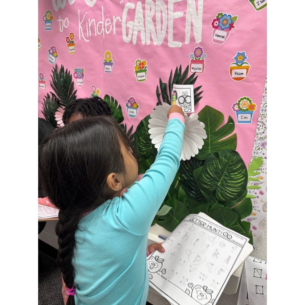 A child in a blue shirt reaches up to add a card to a flower-themed kindergarten welcome board.