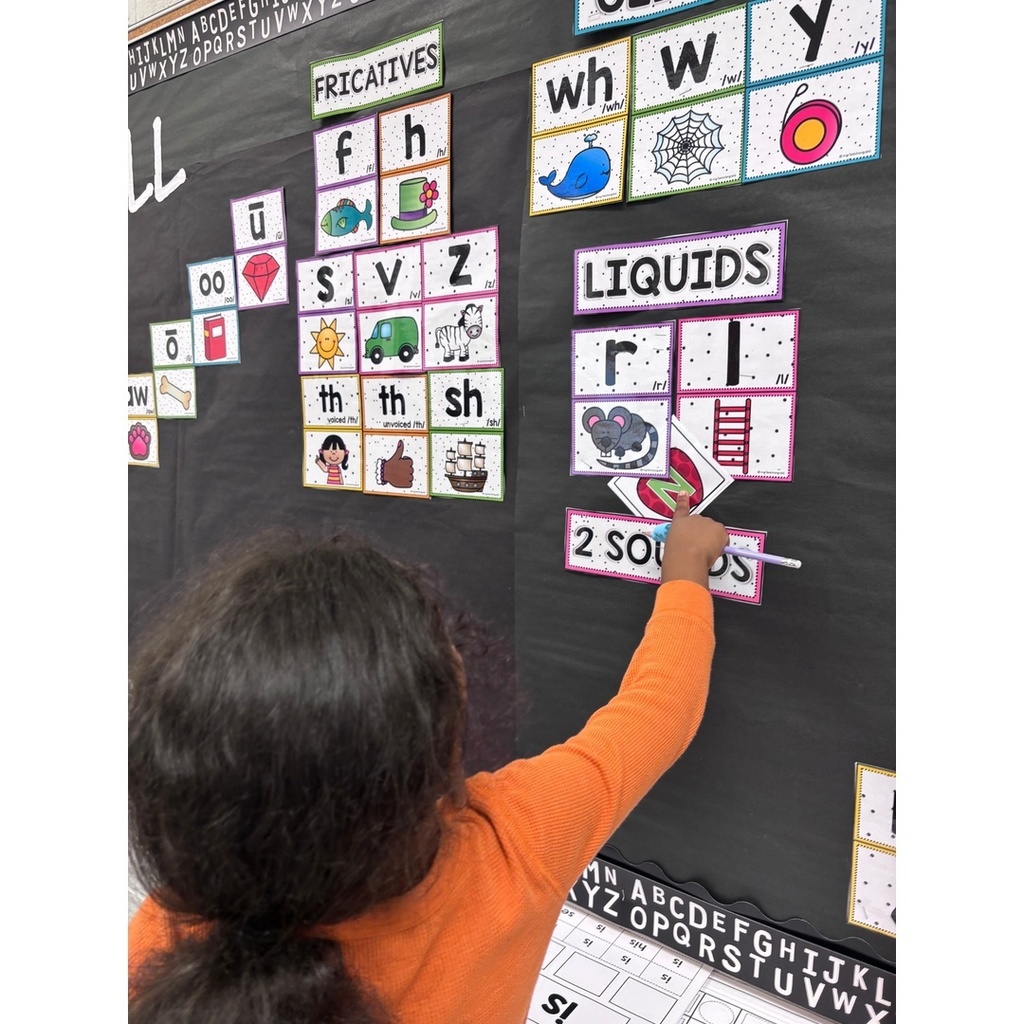 A child in an orange shirt places a letter card onto a wall chart showing sounds.