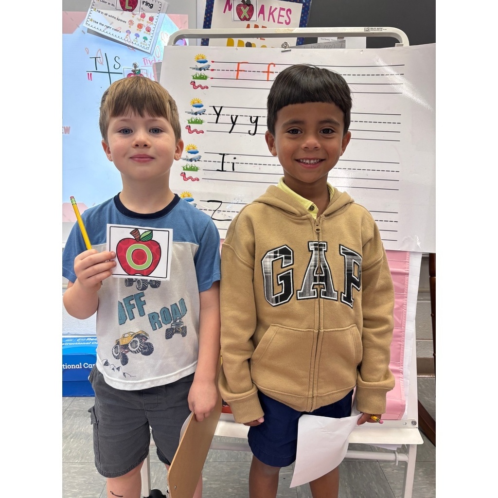 Two students smile at the camera, one holding a letter card and a clipboard, standing in front of handwriting charts.