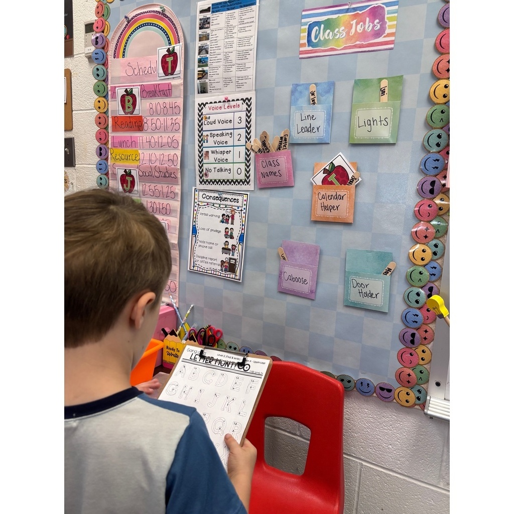 A child looks at a clipboard while standing near a wall with a class jobs chart and schedule.