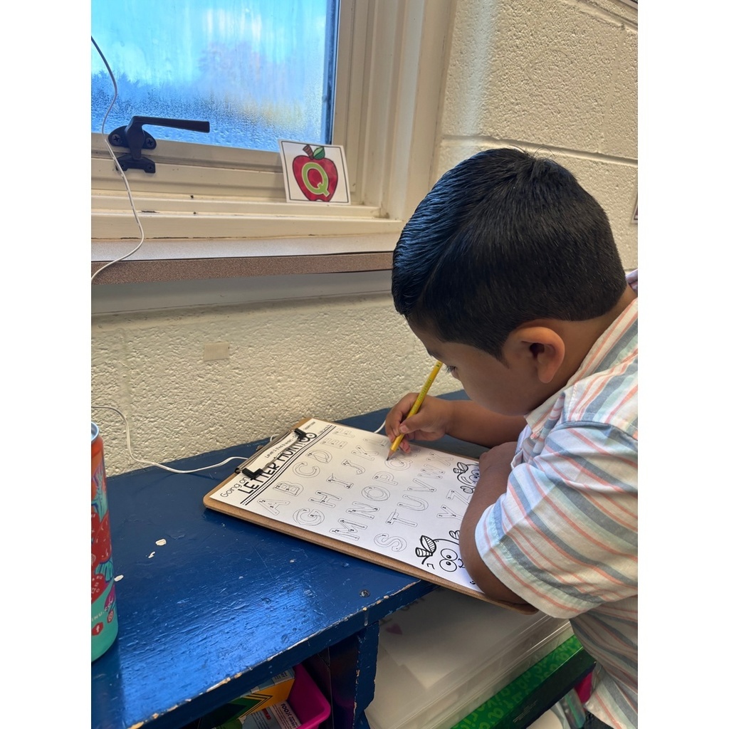 A child works on a letter hunt worksheet at a windowsill with an alphabet card taped nearby.