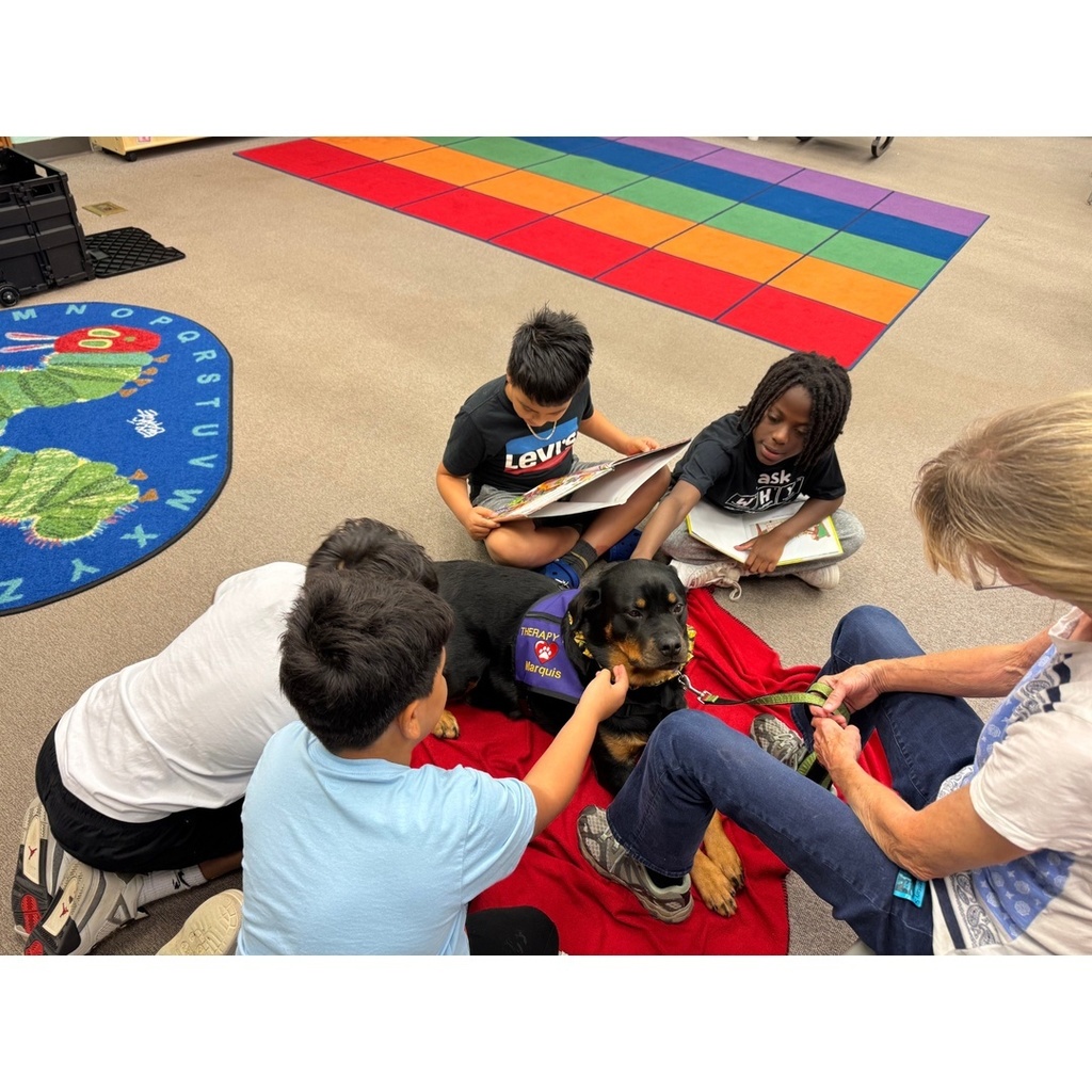 Children sit on a red blanket petting a therapy dog while reading books, guided by an adult.