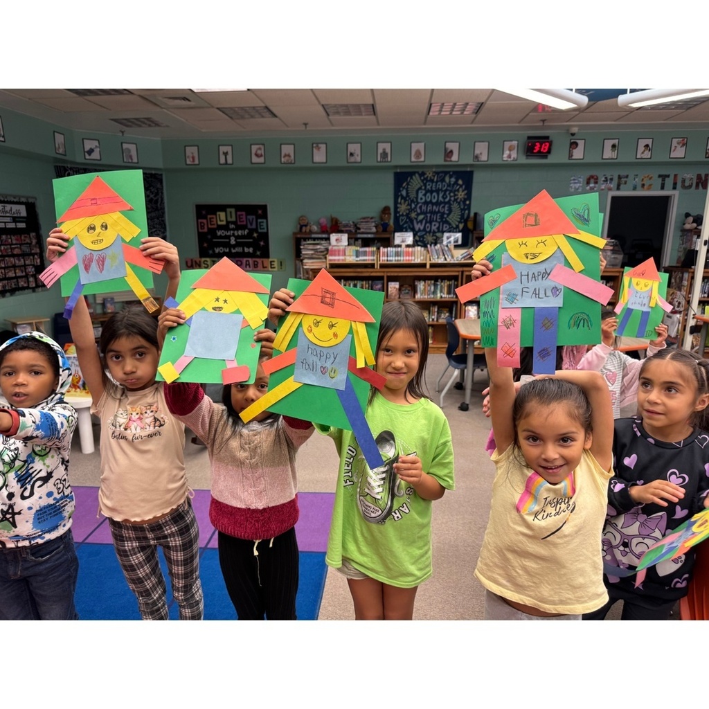 Students smile and hold up bright scarecrow-themed fall crafts with “Happy Fall” messages.
